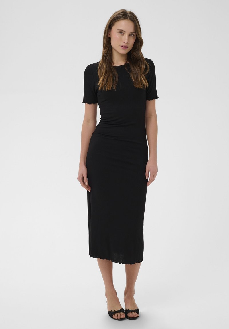 Jeune femme debout portant une robe midi noire ajustée à manches courtes et des talons ouverts noirs, devant un fond clair uni.