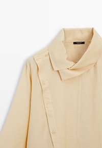 Beige overhemd met knopen en een brede kraag, sluit aan de voorkant met knopen, lange mouwen en een zacht, gestructureerd stof. Merklipje zichtbaar.