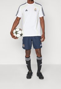 Camiseta de fútbol blanca de Adidas con detalles verdes, pantalones cortos azul oscuro, medias negras con rayas blancas y botas de fútbol negras, sosteniendo un balón de fútbol.