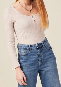 Maglietta a maniche lunghe a coste di un rosa chiaro con scollatura a scalloped e tre bottoni, abbinata a jeans blu in denim a vita alta.