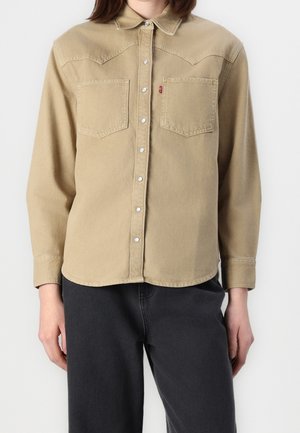 Femme portant une chemise beige à boutons-pression avec des poches poitrine, associée à un pantalon noir, se tenant devant un arrière-plan blanc uni.