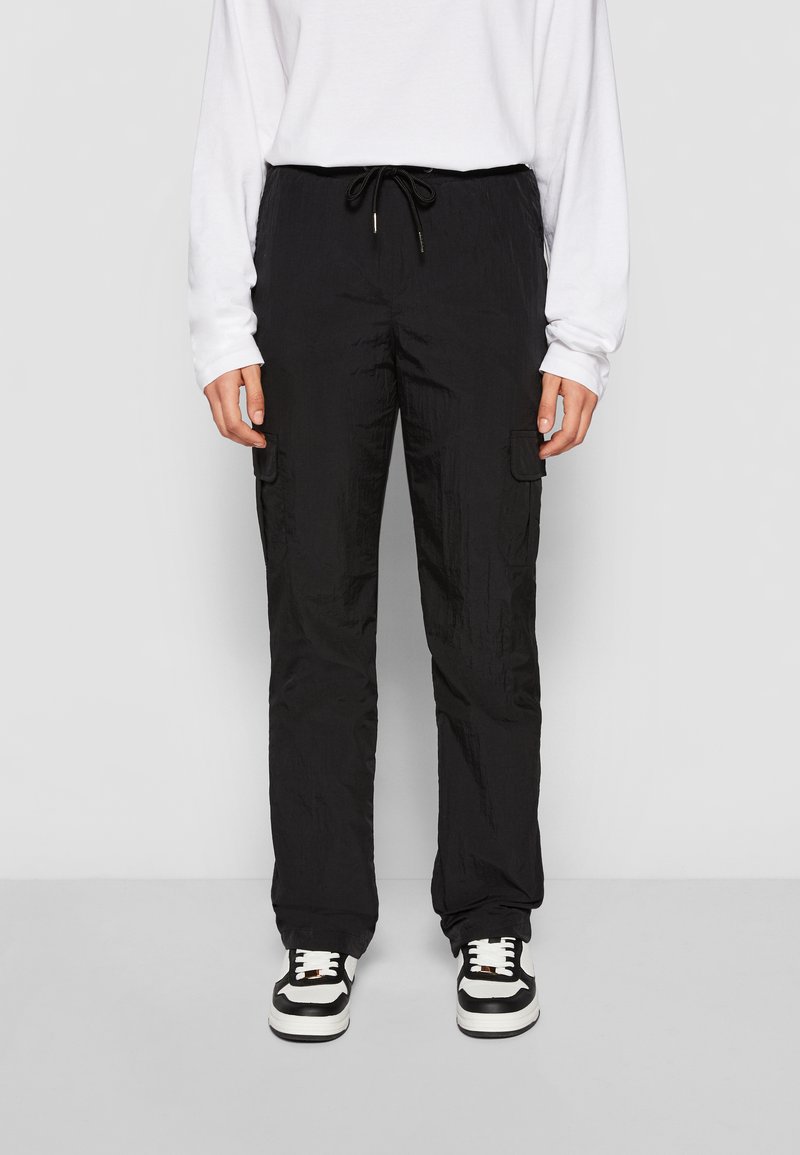 Personne portant un pantalon cargo noir avec des poches latérales, une chemise blanche à manches longues et des baskets noir et blanc se tenant contre un fond uni.