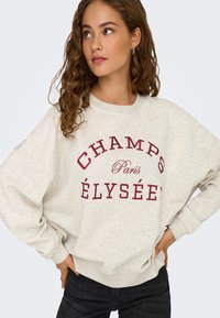 Ljusgrå oversize sweatshirt med texten "CHAMPS ÉLYSÉE" i vinröd färg. Mjuk textur med ribbade ärmslut och midja.