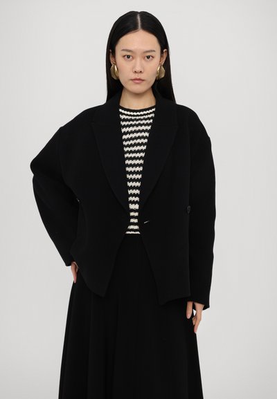 Schwarzer oversized Blazer mit breiten Revers und einem einzelnen Knopfverschluss, getragen über einem schwarz-weiß gestreiften Pullover. Glatte Textur.
