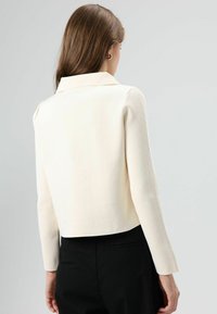 Veste structurée de couleur crème avec un col et des manches longues, présentant un tissu texturé et un coupe courte, portée sur un pantalon noir.