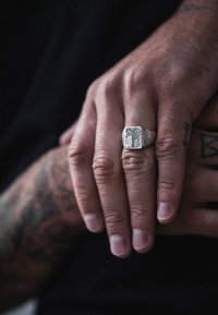 Sidabrinis žiedas su raižytu palmės medžio dizainu, kvadratinės formos ir tekstūruotu žiedu, ilsintis ant rankų su matomais tatuiruotėmis.