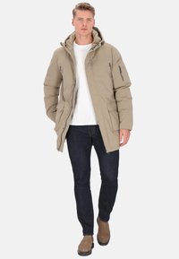 Giacca imbottita beige con cappuccio, tasche con zip e vestibilità comoda, abbinata a jeans scuri e stivali marroni. Texture liscia.