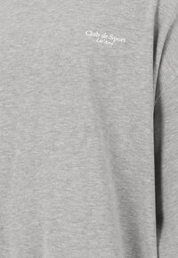 Grå långärmad skjorta i mjukt material. Har en vit logotyp med texten "Club de Sport Les Arcs" på vänster bröstområde.