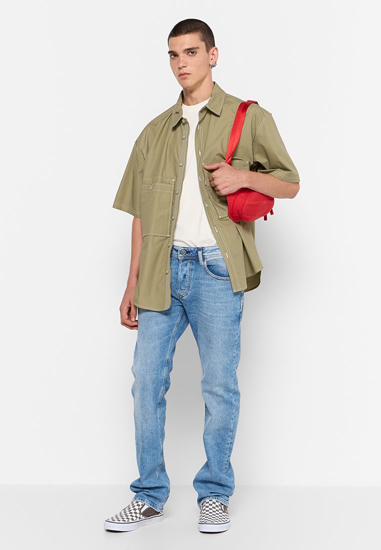 Giovane uomo che indossa una camicia khaki a maniche corte, una t-shirt bianca, jeans blu e scarpe slip-on a scacchi, mentre tiene una borsa rossa a spalla.