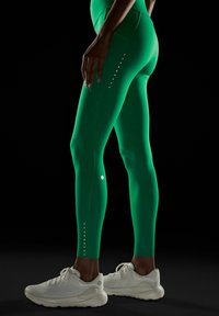 Zeleni visok pas Leggins iz raztegljivega materiala, okrašeni s reflektivnimi pikami ob strani; v kombinaciji z belimi športnimi čevlji.