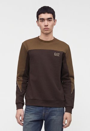 Jeune homme portant un sweat-shirt marron et vert olive foncé à col rond avec le logo EA7 sur la poitrine, associé à un jean bleu, debout devant un fond uni.