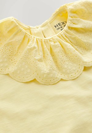 Žlutý top s nařaseným límečkem zakončeným vyšívaným vroubkovaným lemem. Vyrobený z měkké látky, má hladkou texturu a jednoduchý design.