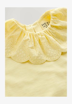 Žuta bluza s naboranim ovratnikom i izvezenim nazubljenim rubom. Izrađena od mekanog materijala, ima glatku teksturu i jednostavan dizajn.