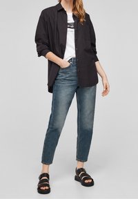 Svart knappad skjorta, vit grafisk t-shirt under, blå jeans och svarta sandaler. Avslappnad passform med en lös silhuett och uppkikade ärmar.