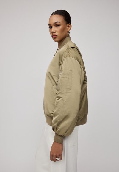 Khaki bomberka z prążkowanymi mankietami i dołem. Posiada kieszenie zapinane na zamek oraz gładką, błyszczącą teksturę. Przedstawiona w profilu bocznym.