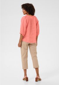 Top in lino corallo con maniche a palloncino e scollatura volant, abbinato a pantaloni cropped beige chiaro e sandali con tacco a blocco marroni.