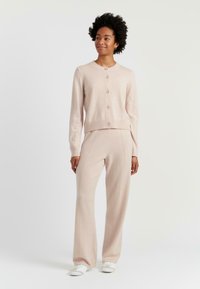 Cardigan tricoté rose clair, à col rond et six boutons, associé à un pantalon large rose clair assorti. Chaussures blanches.