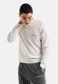 Maglione bianco a maniche lunghe con scollo rotondo, dotato di un logo ricamato nero sul petto. Abbinato a pantaloni grigio scuro, entrambi dal look casual.