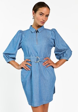 Vrouw draagt een blauw gestreepte jurk met ceintuur, met opgeblazen driekwartmouwen en een ritskraag, poseert met handen in haar zij.