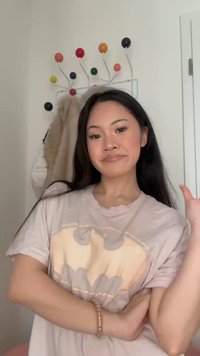 Junge Frau mit langen dunklen Haaren, die ein helles T-Shirt mit Batman-Logo trägt und mit verschränkten Armen vor einem bunten, an der Wand montierten Garderobenständer steht.