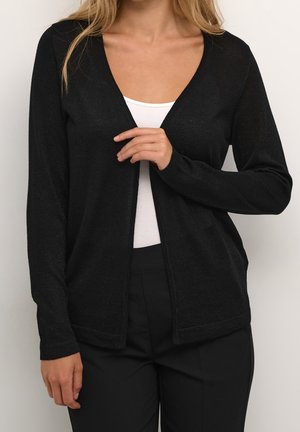 Femme portant un cardigan noir ouvert sur un haut blanc et un pantalon noir, vue des épaules aux hanches, ajustant le cardigan d'une main.