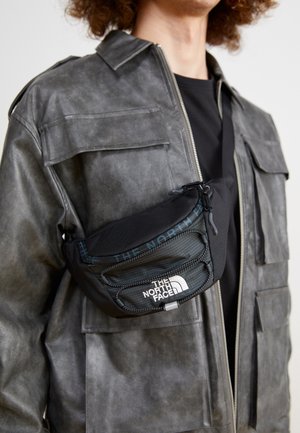 Osoba nosząca szarą skórzaną kurtkę i czarną torbę na ramię z logo The North Face na przedniej kieszeni.