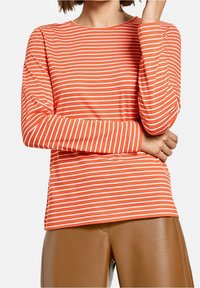 Langärmliges, Rundhalsoberteil in leuchtendem Orange mit weißen horizontalen Streifen, aus weichem Stoff, kombiniert mit braunen Kunstlederhosen.