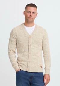 Mann med kort hår iført beige knappet cardigan over hvit skjorte og blå jeans, står mot en lys bakgrunn.
