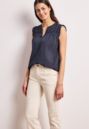 Femme portant un chemisier sans manches bleu marine avec des épaules brodées et un pantalon crème, souriante sur un fond clair uni.