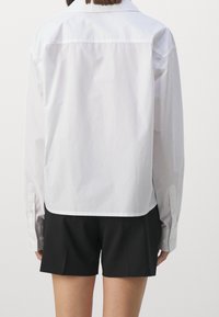 Camicia bianca a maniche lunghe con colletto a punta, polsini abbottonati e una texture liscia, indossata sopra pantaloni corti neri.