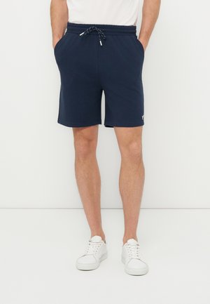 Mężczyzna w granatowych, ściąganych w pasie szortach i białych trampkach, stojący z rękami w kieszeniach na białym tle.