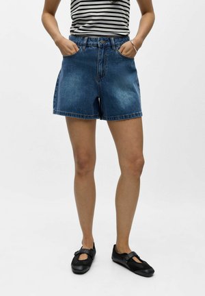 Femme portant un short en denim bleu taille haute, des chaussures noires de style Mary Jane, et un haut sans manches rayé noir et blanc, debout les mains dans les poches.