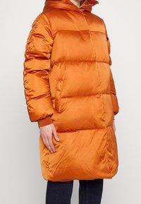 Vadderad orange jacka med huva, med horisontell vaddering, påsydda fickor och ribbstickade muddar. Tillverkad av ett glansigt, slitstarkt tyg.