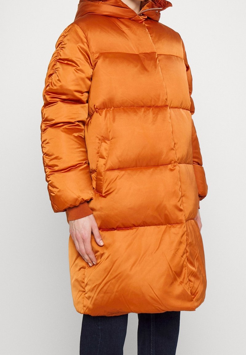 Vadderad orange jacka med huva, med horisontell vaddering, påsydda fickor och ribbstickade muddar. Tillverkad av ett glansigt, slitstarkt tyg.