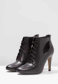 Mustat nahkaiset nilkkurit, joissa on terävä kärki, nyöritys edessä, leikatut sivut ja ohut piikkikorko. Sileä viimeistely, jossa hillitty kiilto.