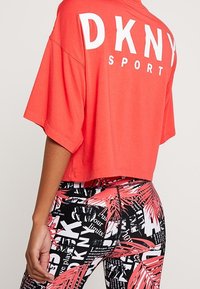 Röd kort t-shirt med "DKNY SPORT" tryckt i vitt på ryggen, kombinerad med svarta leggings med ett mönster av flerfärgade palmblad och text.