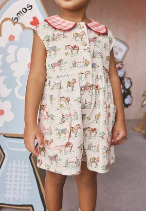 Kind trägt ein ärmelloses Kleid mit pinkfarbenem kariertem Kragen und Pferde- und Stallmuster, steht drinnen in der Nähe von bunter Wandkunst.