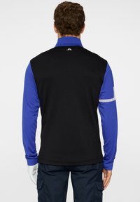 Svart och blå långärmad väst med texturerad baksida, framkrage, vit randig accent på ärmen och logodetalj.