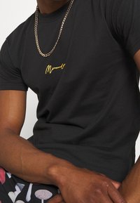 T-shirt in cotone nero con testo ricamato giallo "Memories" sul petto, abbinato a una collana di catena in argento.