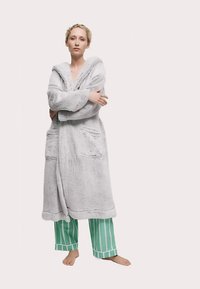 Grå faux pelsbadekåbe med hætte, foran bindebånd og lommer, båret over grønne og hvide stribede pyjamasbukser. Blød tekstur og løs pasform.