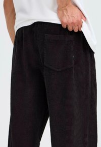 Schwarze Cordhosen verfügen über vertikale Rillen, eine Gesäßtasche und eine lockere Passform. Der Stoff hat eine weiche Textur, die Komfort und Wärme bietet.