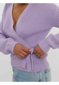 Liliowa dzianinowa kardigan z głębokim dekoltem w kształcie V, długimi rękawami i związanym w pasie, z wyraźnym wzorem. Noszony na jasnoniebieskich dżinsach.