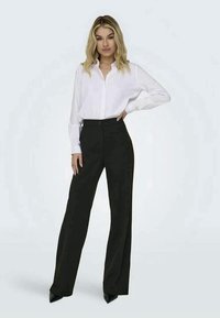 Chemise blanche à boutons associée à un pantalon large noir, tous deux en tissu lisse. Les pantalons présentent une fermeture à bouton à l'avant et des poches en biais.