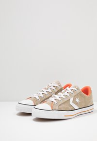 Beżowe płótniowe trampki z białą gumową podeszwą, pomarańczowymi akcentami i białym logo gwiazdy. Posiadają sznurowany design i kontrastowe tekstury.