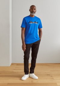 T-shirt in cotone blu con testo grafico nero, abbinata a pantaloni neri e sneakers bianche. Il modello è in piedi contro uno sfondo neutro.