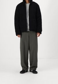 Veste en polaire noire texturée avec fermeture éclair, deux poches avant, associée à un pull rayé gris clair et à un pantalon vert olive.