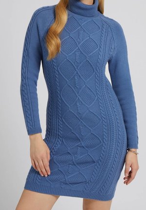 Femme portant une robe-pull bleu ajustée à col roulé en maille torsadée, avec manches longues et motifs diamant texturés.