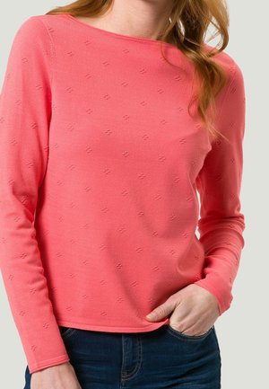Pull à manches longues en corail vif, avec un motif texturé et un col rond, associé à un jean bleu foncé.