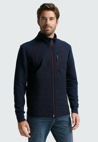 Navyblaue Zip-Jacke mit gesteppter Vorderseite, hohem Kragen und rotem Akzent-Reißverschluss. Sie hat lange Ärmel und ein schlankes Design.