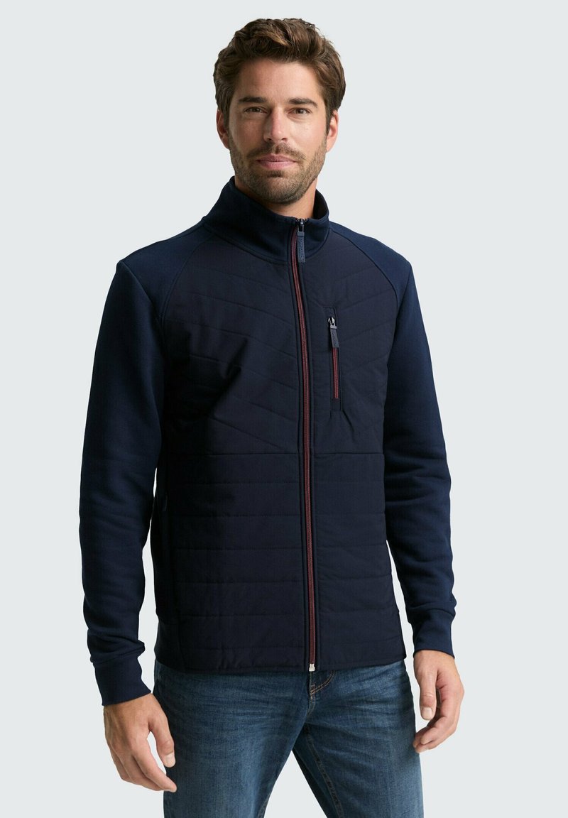 Navyblaue Zip-Jacke mit gesteppter Vorderseite, hohem Kragen und rotem Akzent-Reißverschluss. Sie hat lange Ärmel und ein schlankes Design.